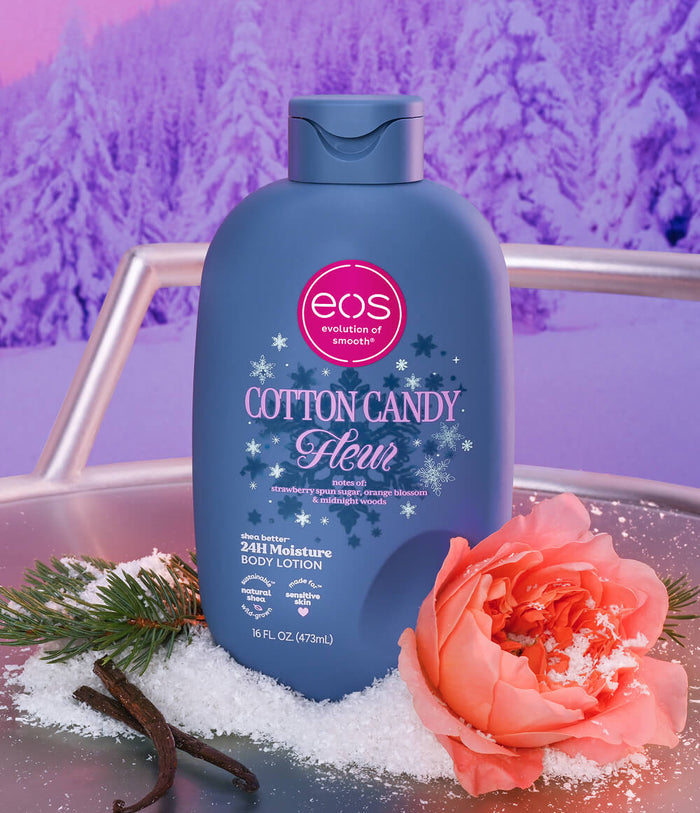 A bottle of EOS Cotton Candy Clean body lotion stands on a reflective surface with decorative winter-themed props, including a peach rose, snow, and vanilla beans, against a purple snowy forest backdrop.