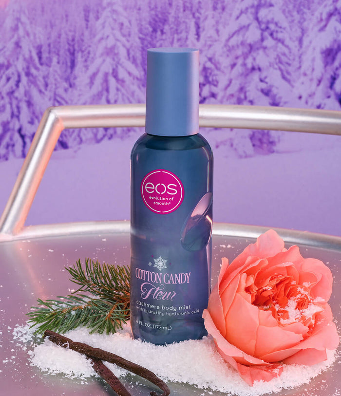 A bottle of EOS Cotton Candy Fleur glow body mist stands on a reflective silver tray surrounded by white sugar, a peach rose, and cinnamon sticks, with a purple textured background.