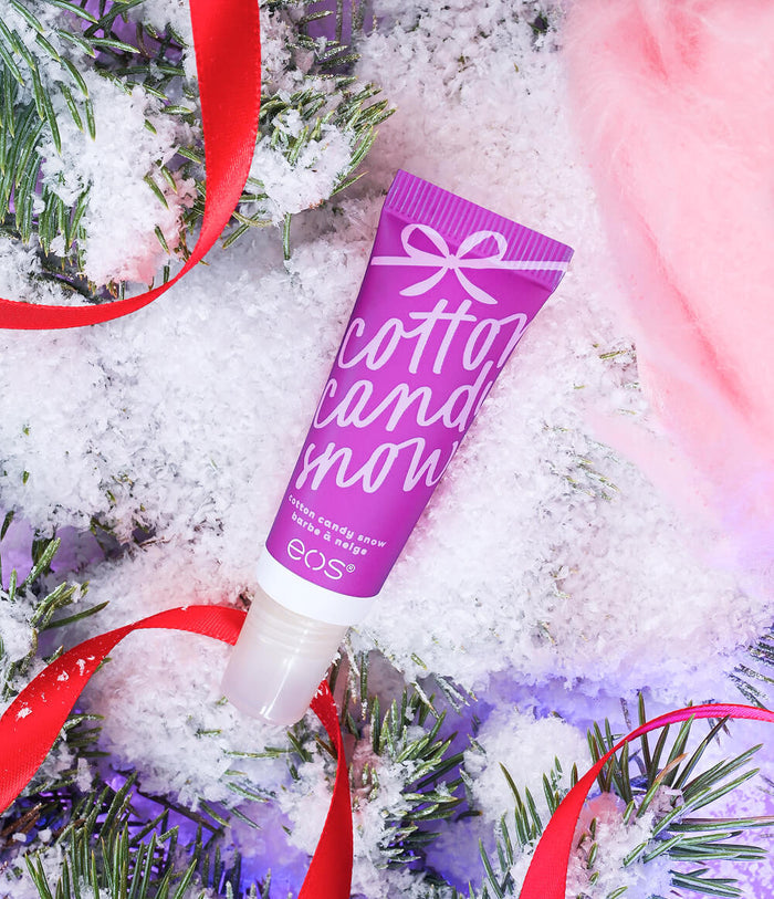 A purple squeeze tube of EOS Cotton Candy Snow lip balm is styled in a winter holiday flat-lay with snow, pine branches, pink candy cane decorations, and red ribbon.