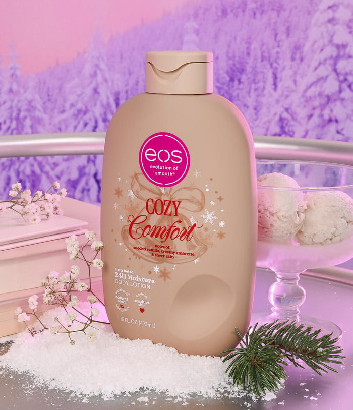 A bottle of EOS Cozy Comfort body lotion stands on a tabletop styled with faux snow, baby's breath, a pine sprig, and a glass of ice cream, against a pink and purple winter landscape background.