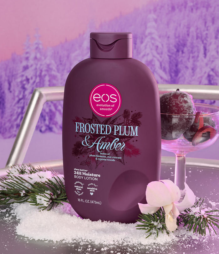A bottle of EOS Frosted Plum & Amber body lotion is displayed in a festive winter setting with artificial snow, pine branches, a white orchid, and a cocktail glass with berries against a purple winter forest backdrop.