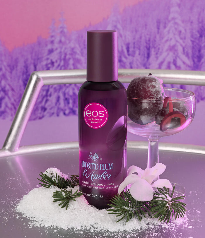 A bottle of EOS 'Frosted Plum & Amber' glacial body mist sits on a reflective tray next to a glass bowl of frosted plums, with white sugar crystals and green pine sprigs in the foreground against a purple winter forest backdrop.