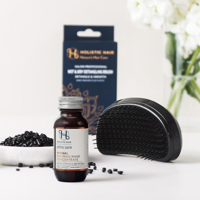 Holistic Hair Herbal Detangling Rinse bottle with its packaging box, a black detangling brush, and scattered black hair caps on a white surface with white flowers in the background.