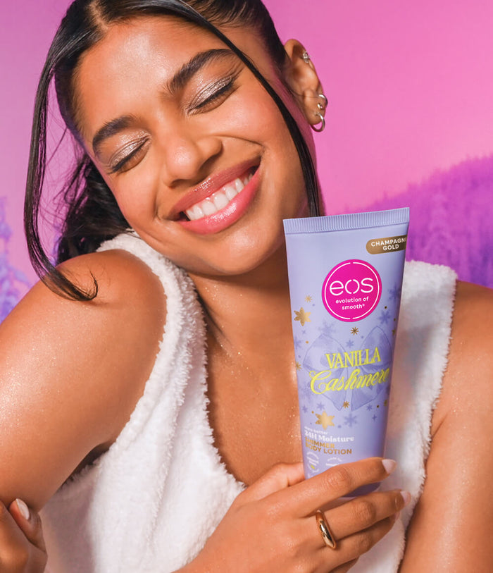 A smiling woman in a white towel robe holds a tube of EOS Vanilla Cashmere Shimmer body lotion against a vibrant pink background.