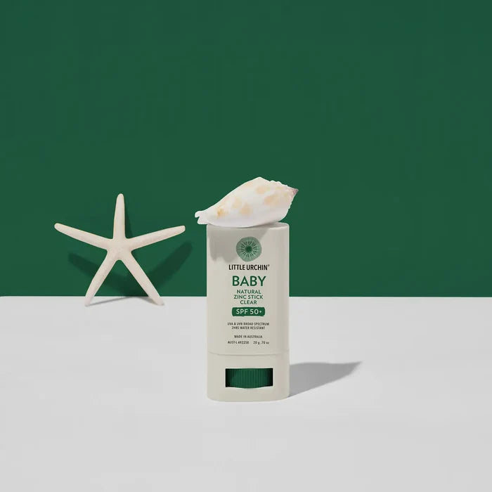 A baby sunscreen stick on a white surface against a dark green background, styled with a white seashell on top of the stick and a white starfish prop on the left.
