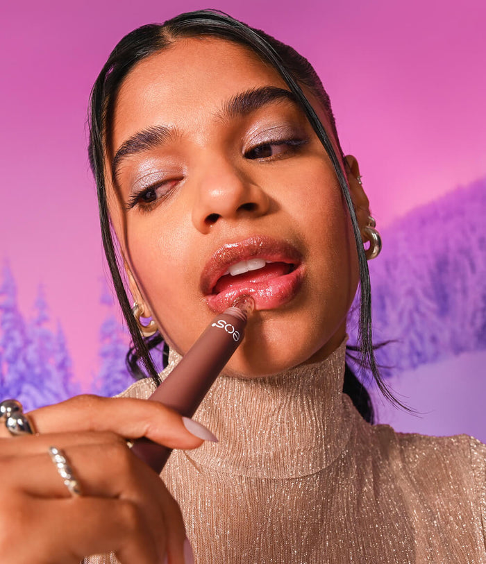 A close-up lifestyle shot of a woman applying a lip gloss. She is wearing a sparkly beige turtleneck and gold hoop earrings, set against a vibrant pink background with a stylized purple winter forest silhouette.