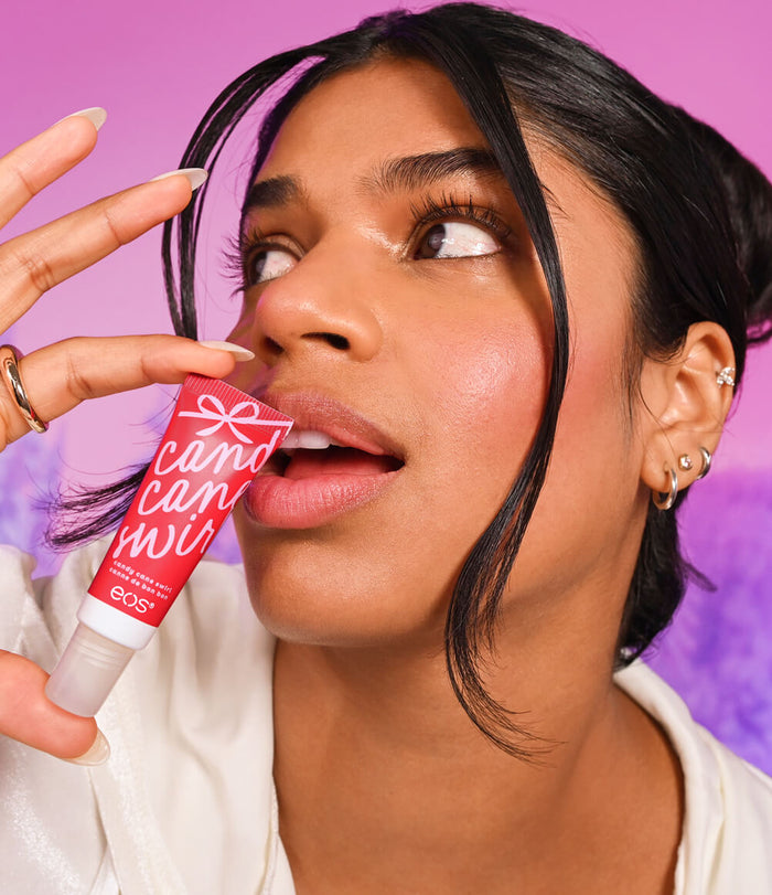 A close-up lifestyle shot of a woman holding a red tube of EOS Candy Can Melt lip balm near her lips, set against a vibrant pink and purple gradient background.