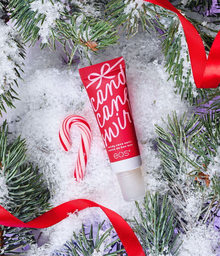 A red tube of EOS Cosmetics 'Candy Cane Wit' lip balm is styled in a festive flat-lay with snow, pine branches, and red ribbons, alongside a candy cane prop.