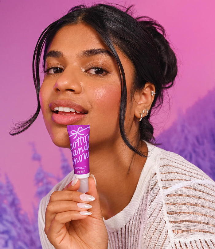 A woman holding a purple tube of  lip balm against a vibrant pink and purple background.