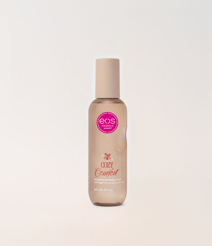 A bottle of EOS Cosmetics Cozy Comfort Body Mist in a frosted beige bottle with a matching beige cap, featuring a pink circular EOS logo on the front, set against a clean off-white background.