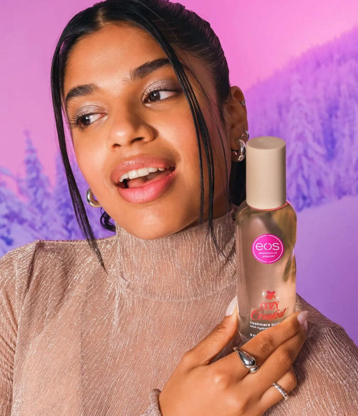 A woman holding a bottle of EOS body spray against a pink and purple winter forest backdrop.