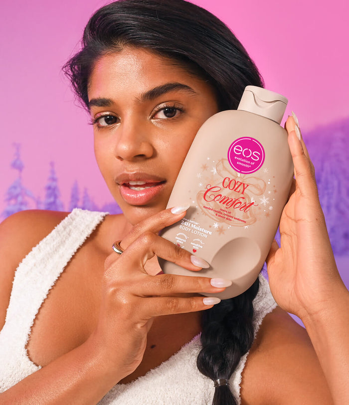 A woman holds a large bottle of EOS Cozy Comfort Hand & Foot Cream against a pink and purple gradient background. She is wearing a white towel top, and the bottle is beige with a pink circular EOS logo.
