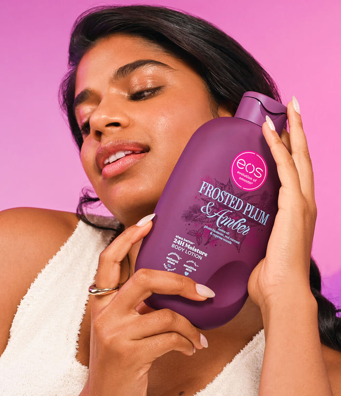 A woman in a white towel holds a large bottle of EOS Frosted Plum & Amber body wash against a solid pink background.