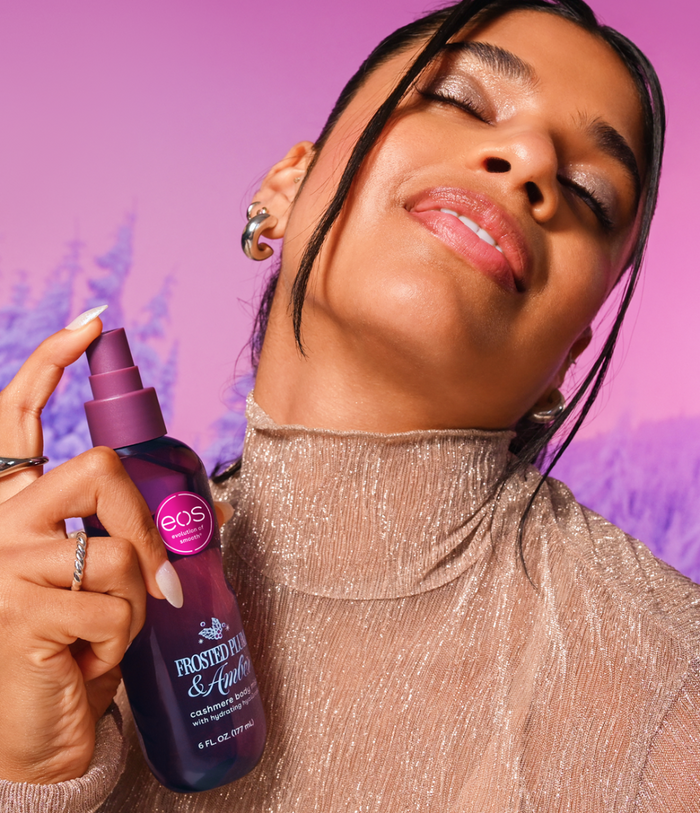 A woman in a shimmering beige turtleneck holds a bottle of EOS against a vibrant pink and purple background.