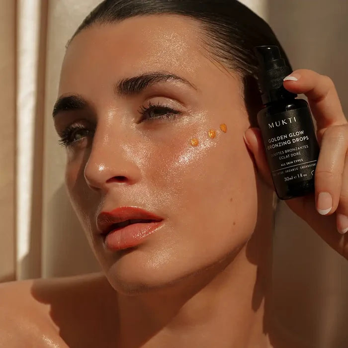 A close-up of a woman holding a black bottle of MUKT Gold Glow Golden Enhancing Drops next to her cheek where three golden drops of serum are applied, showing a glowing, hydrated complexion.