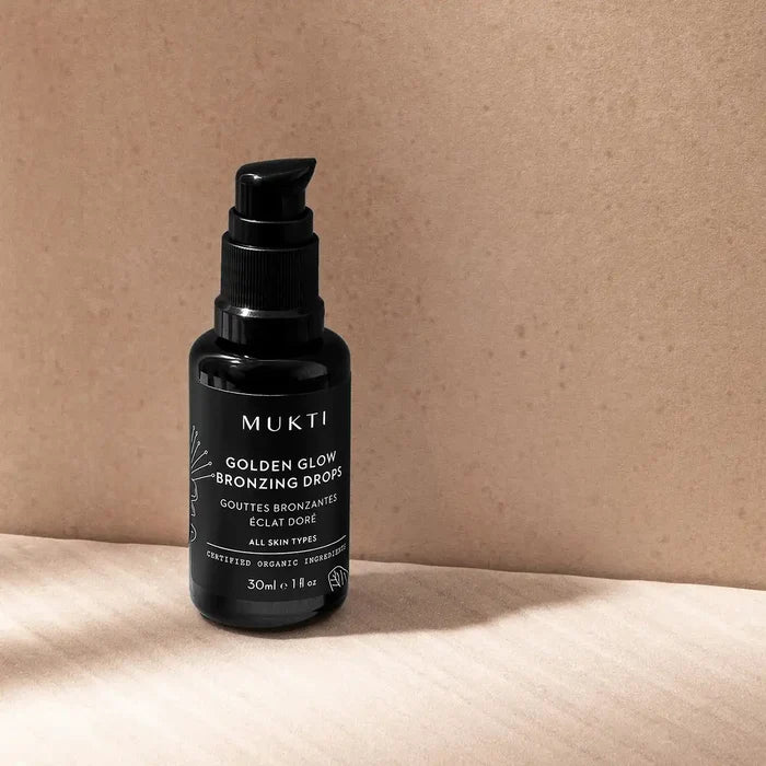 A black pump bottle of MUKTI Golden Glow Bronzing Drops placed on a light wood surface against a textured beige background.