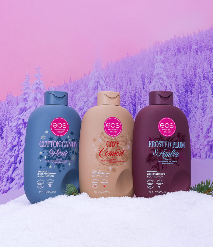 Three EOS Home & Personal Care body lotion bottles standing in white snow against a pink and purple snowy mountain backdrop. The bottles are labeled Cotton Candy Glaze in blue, Cozy Comfort in beige, and Frosted Plum & Amber in dark plum.