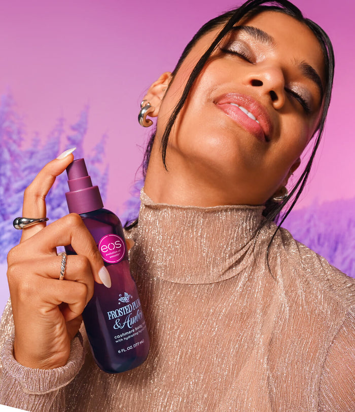 A woman with closed eyes holds a bottle of EOS body spray against a pink and purple background.