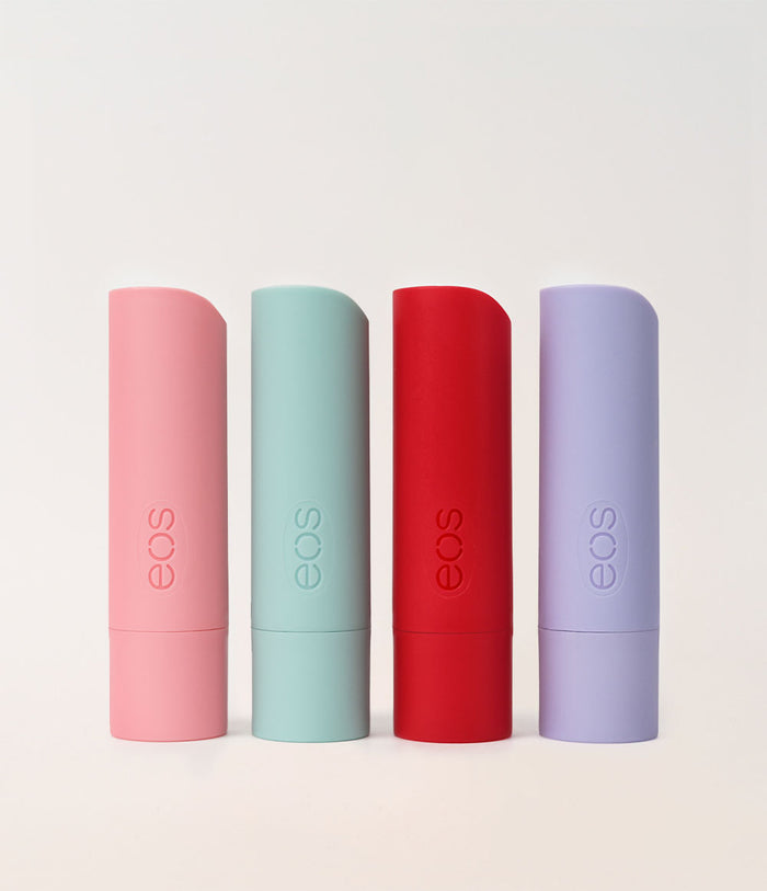 Four EOS lip balm tubes in pastel pink, mint green, red, and lavender purple, standing upright in a row against a light off-white background.