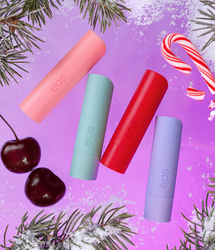 Four EOS lip balm tubes in pastel pink, mint green, red, and lavender purple, arranged diagonally on a purple background with Christmas-themed flat-lay props including pine branches, cherries, and candy canes.