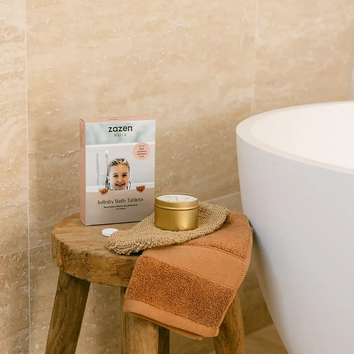 A box of zazen Infinity Bath Tablets sits on a rustic wooden stool next to a white freestanding bathtub, with a terracotta bath towel and a gold tin candle resting on it.