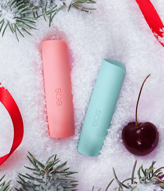 Two EOS lip balm tubes, one in coral pink and one in mint green, placed in white snow with pine branches, a red ribbon, and a dark cherry.