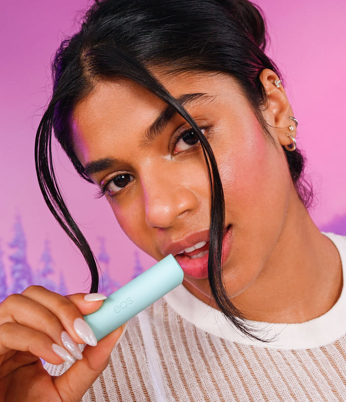 A close-up lifestyle shot of a woman holding a light blue cylindrical beauty product, with the brand name 'bce' visible on the tube. She is wearing a white ribbed knit top against a vibrant pink background.