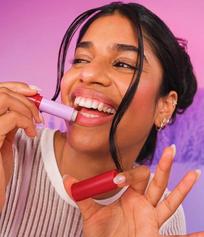A woman with dark hair pulled back smiles while applying a lip product from a red tube with a lavender cap, set against a vibrant pink and purple studio background.