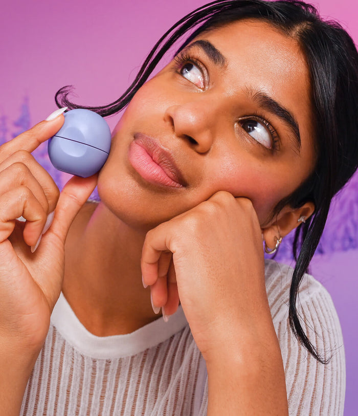 A woman with dark hair looking thoughtfully to the side while holding a small, round, light blue lip balm case between her fingers against a solid purple background.
