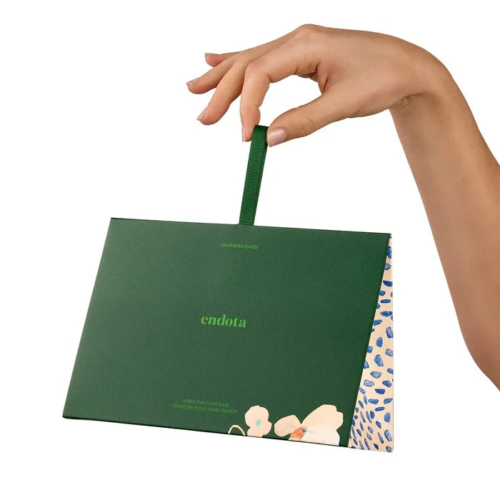 A hand holding a dark green Endota gift set with a matching green handle, shown against a plain white background. The bag features the brand name 'endota' in light green text and a floral illustration near the bottom, with a patterned side panel visible.