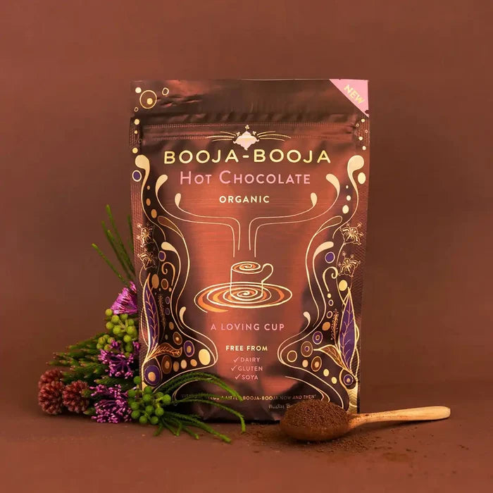 A stand-up pouch of Booja-Booja Hot Chocolate Organic coffee is displayed on a warm brown background, styled with a small wooden spoon of dark chocolate powder and dried flowers on the left.