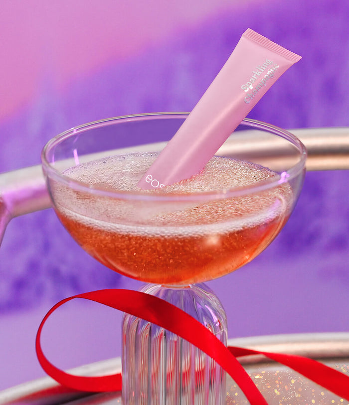 A pink tube of lip shine rests inside a clear glass filled with a sparkling peach-colored drink, tied with a red ribbon against a purple background.