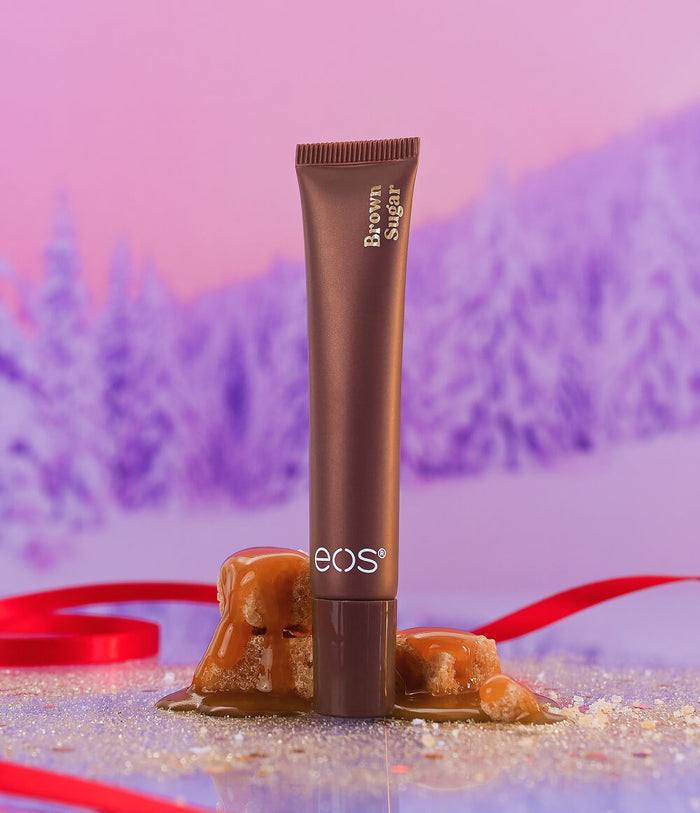 A tube of EOS Brown Sugar cosmetic product stands upright on a glittery surface with caramel drizzle and sugar crystals in the foreground, against a pink and purple blurred winter forest background with a red ribbon.