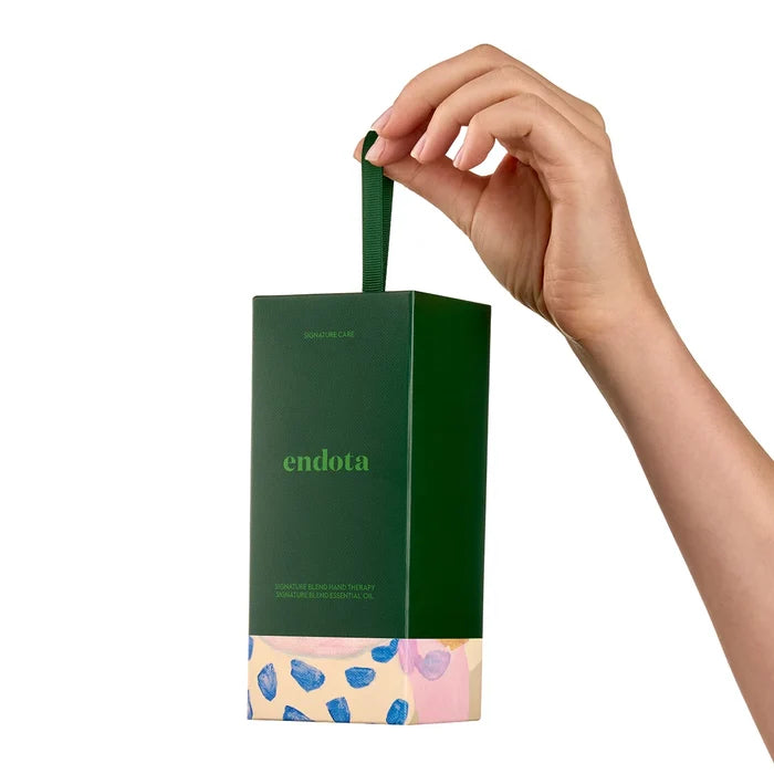 A hand holds a dark green Endota skincare box with a matching green handle against a clean white background. The box features the brand name 'endota' in green and a decorative bottom section with pink, blue, and beige patterns.