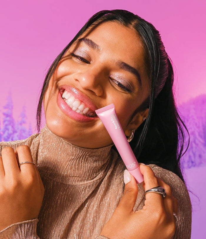 A smiling woman with dark hair holds a pink lip product tube near her mouth, wearing a shiny taupe turtleneck against a pink and purple gradient background.