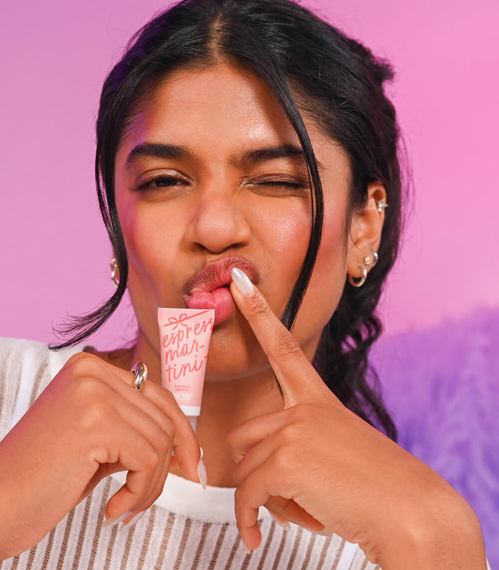 A woman with dark hair and hoop earrings poses with a pink tube of lip balm, licking the packaging while pointing at it with her other hand against a pink and purple gradient background.