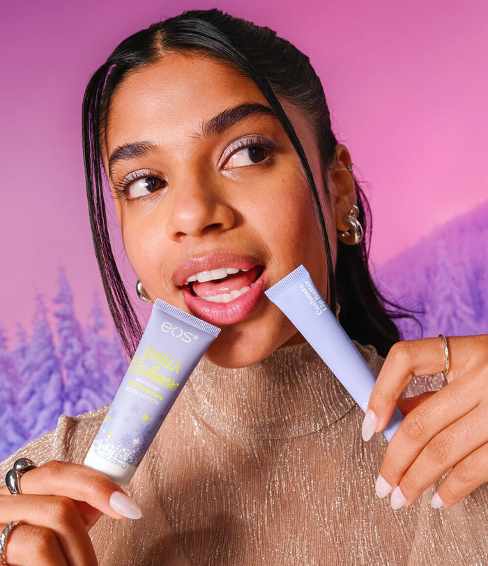 A woman holding two light purple cosmetic tubes, one labeled EOS and the other partially visible, against a pink and purple background with a blurred winter tree scene.